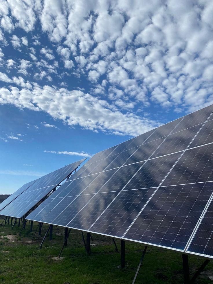 Instalacja fotowoltaiczna z widocznymi panelami na tle błękitnego nieba z chmurami. Panele ustawione pod kątem, odbijają niebo. Widoczne mocowania paneli.