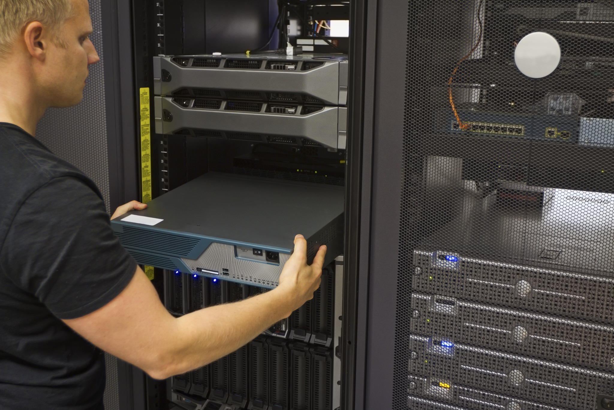 Mężczyzna instaluje serwer Cisco w szafie rack. Widoczne kable, przełączniki sieciowe i inne serwery. Oświetlenie techniczne, metalowa konstrukcja szafy.
