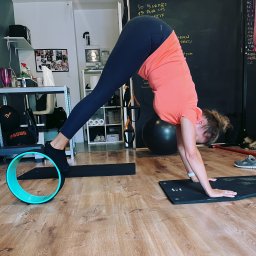 ADRIANA CHRIST REAL FIT - Kobieta w pozycji psa z głową w dół, używa koła do jogi. Ćwiczenie na macie w domowej siłowni, w tle tablica z zapisanymi ćwiczeniami. Trening z akcesoriami.