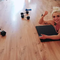 Uśmiechnięta blondynka na macie, uniesiona pięść, tatuaż z hantlami. Wokół hantle, obciążenia. Jasna podłoga, sportowy duch. Trening personalny w domowym zaciszu.