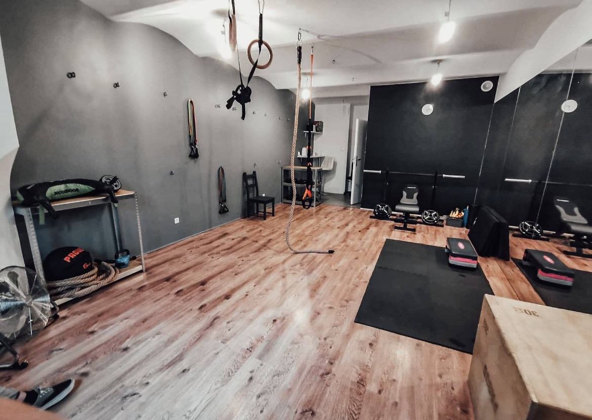 Jasne, przestronne studio treningowe z drewnianą podłogą, wyposażone w sprzęt do ćwiczeń, w tym liny, drabinki i ciężary. Lustrzana ściana.