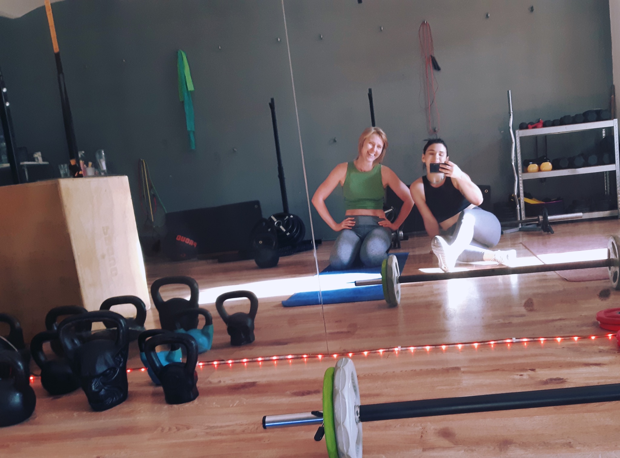 Dwie uśmiechnięte kobiety w sportowych strojach pozują w siłowni, jedna robi selfie. W tle kettlebells, sztanga i drabinki. Widoczne odbicie w lustrze.