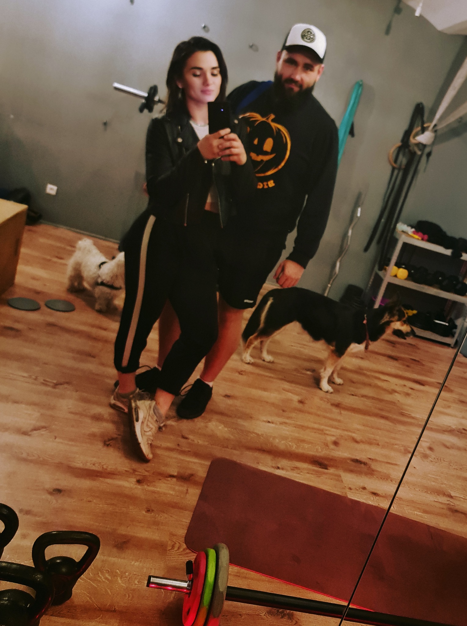 Para robi selfie w domowej siłowni z psami. Widoczne hantle, sztanga, mata do ćwiczeń i regał z obciążeniami. Mężczyzna w bluzie z dynią.