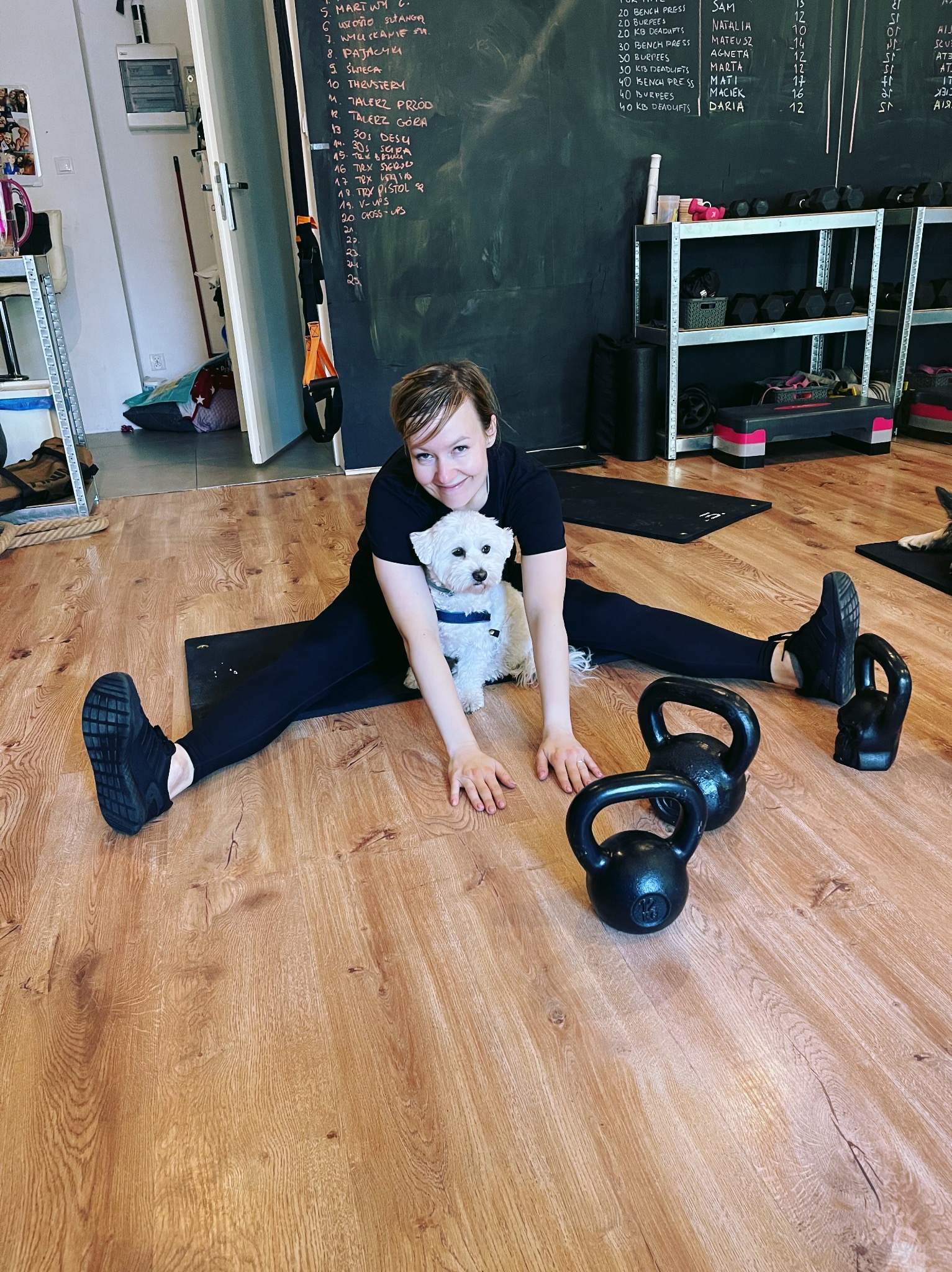 Kobieta w rozkroku na macie, z psem i kettlebells. Tablica z ćwiczeniami w tle. Trening personalny w domowej siłowni. Parkiet z jasnego drewna.