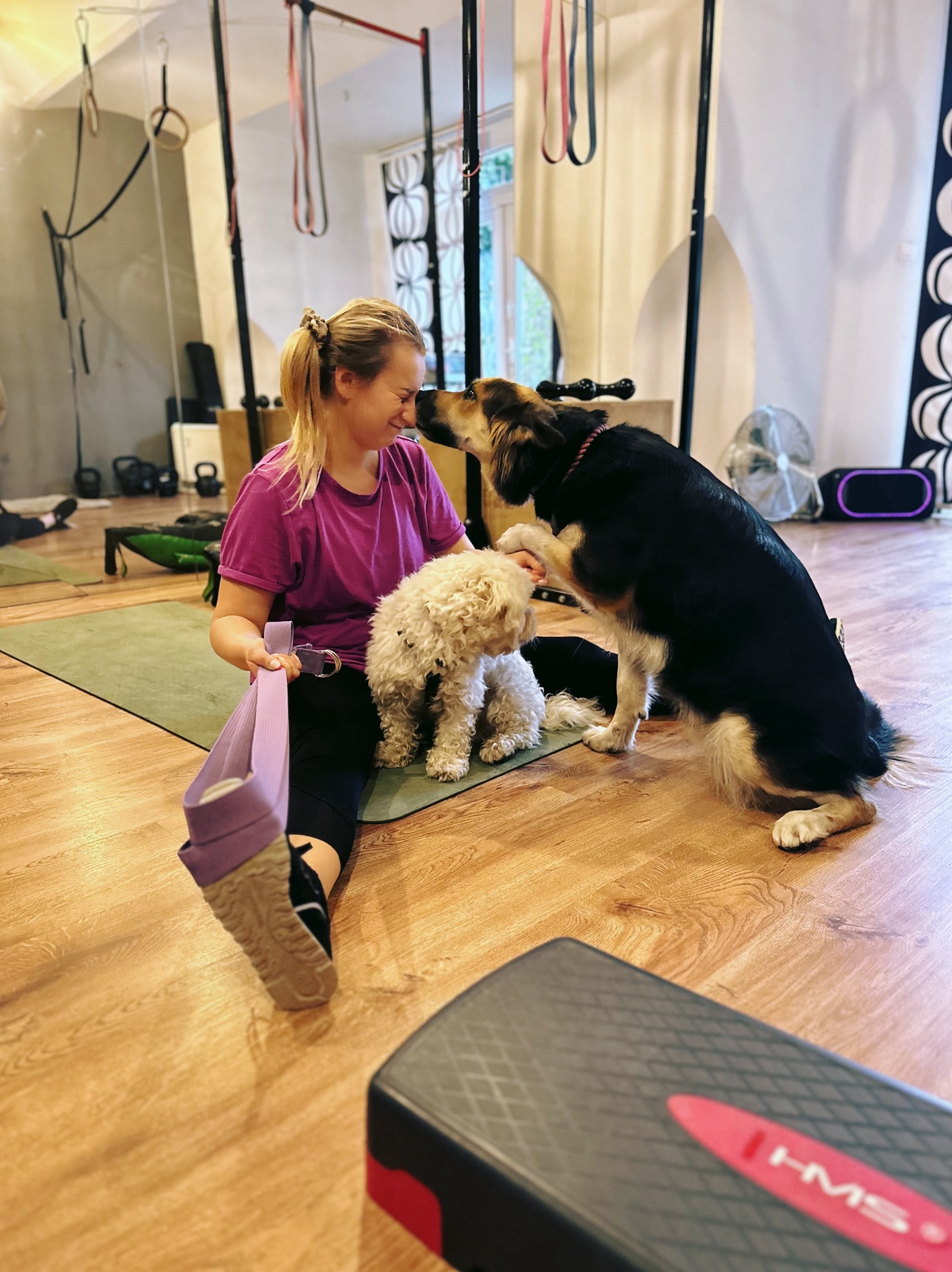 Uśmiechnięta kobieta siedzi na macie w siłowni, trzyma pas do ćwiczeń. Dwa psy, pudel i owczarek, ocierają się o jej twarz. W tle sprzęt do ćwiczeń.