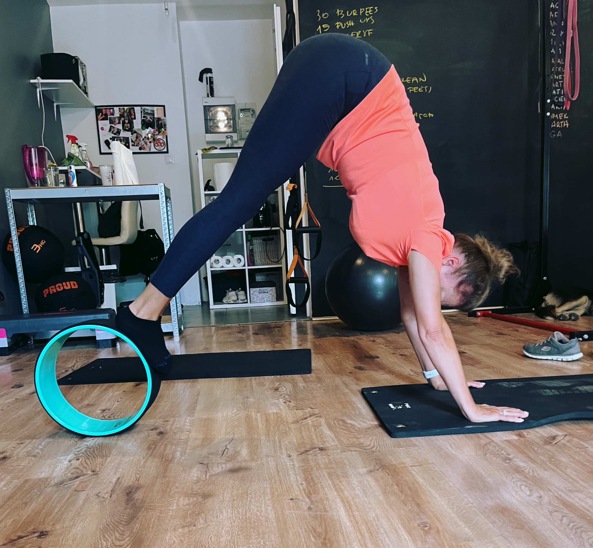 Kobieta w pozycji psa z głową w dół, używa koła do jogi. Ćwiczenie na macie w domowej siłowni, w tle tablica z zapisanymi ćwiczeniami. Trening z akcesoriami.