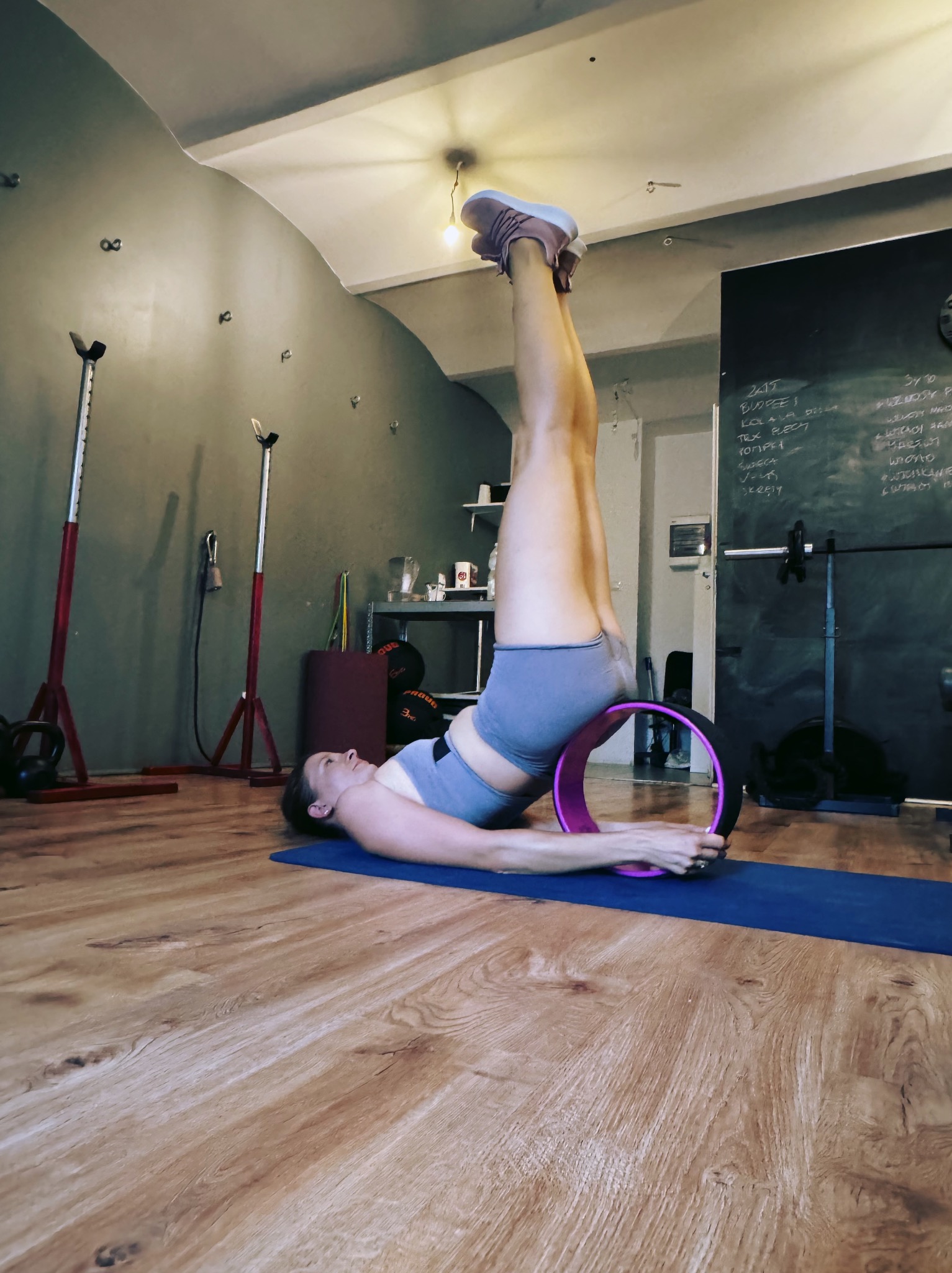 Kobieta ćwiczy z kołem do jogi na macie we Wrocławiu. Widoczne przybory do ćwiczeń i tablica z notatkami. Nietypowy kadr z uniesionymi nogami.