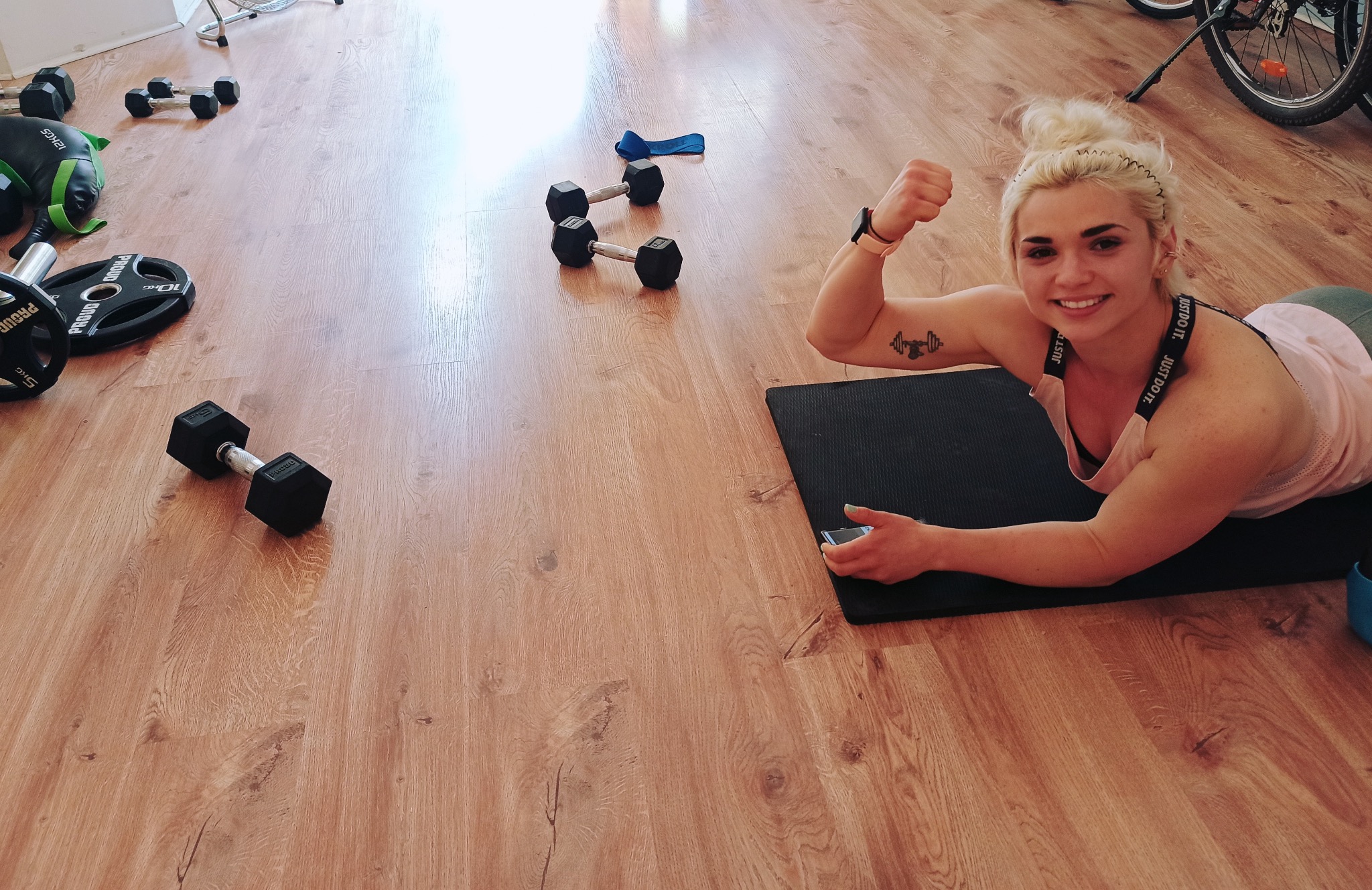 Uśmiechnięta blondynka na macie, uniesiona pięść, tatuaż z hantlami. Wokół hantle, obciążenia. Jasna podłoga, sportowy duch. Trening personalny w domowym zaciszu.