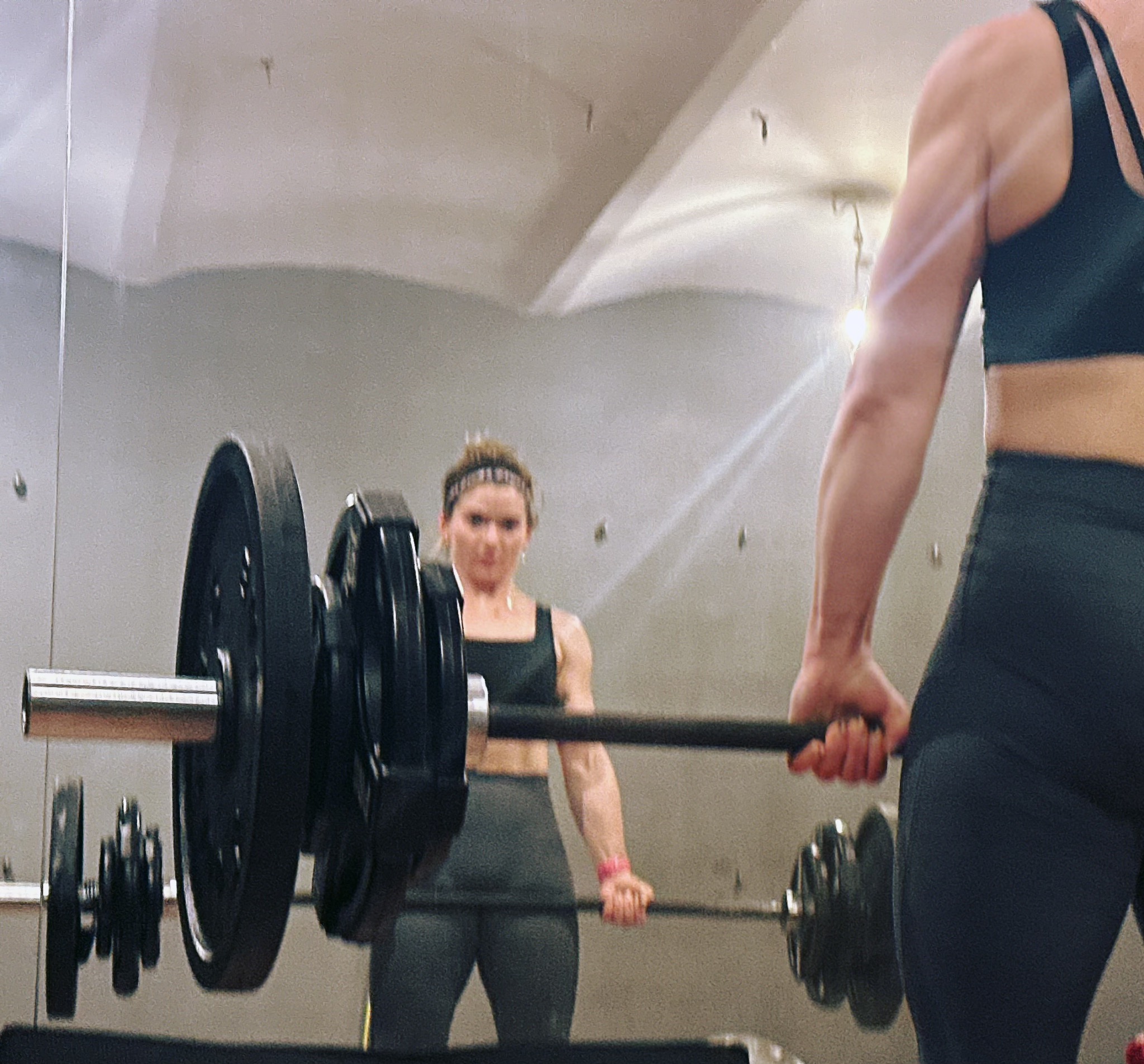 Kobieta w sportowym stroju, odbicie w lustrze podczas podnoszenia sztangi na siłowni. Widoczne obciążenia, gryf i fragment sylwetki drugiej osoby.