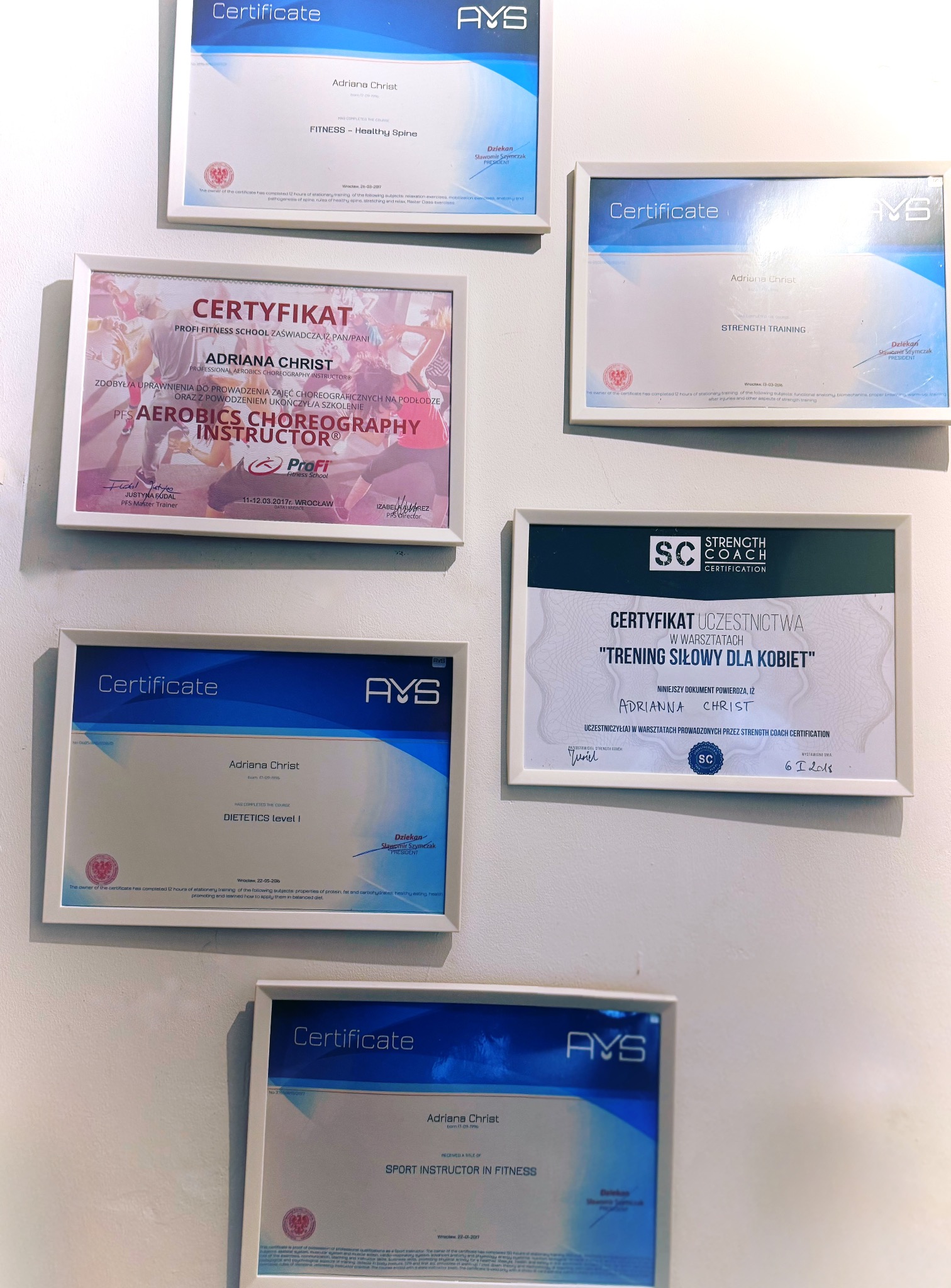Ściana z ramkami prezentującymi certyfikaty i uprawnienia trenerki fitness Adriany Christ z zakresu aerobiku, dietetyki, treningu siłowego i zdrowego kręgosłupa.