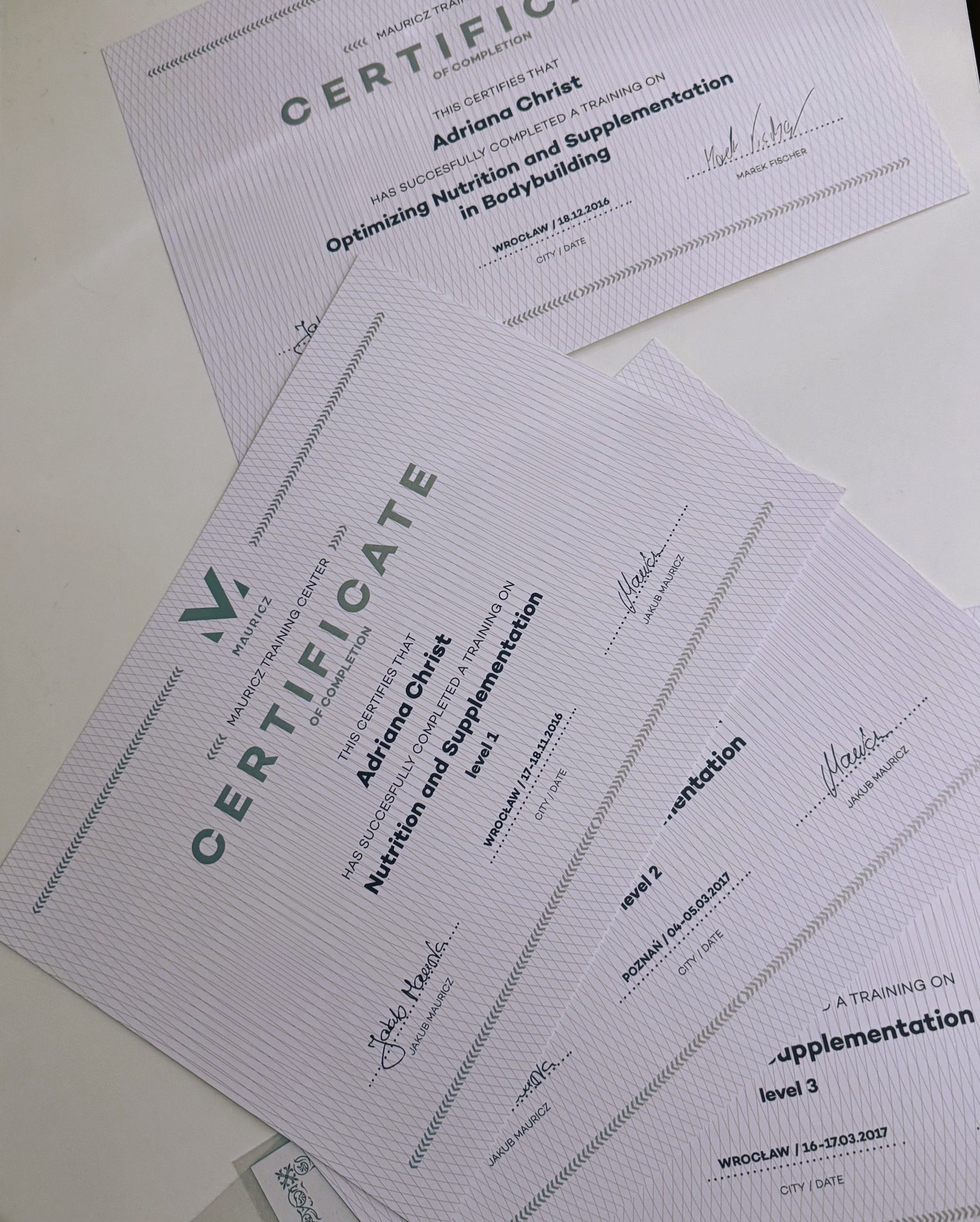 Certyfikaty ukończenia szkoleń z zakresu optymalizacji żywienia i suplementacji w sportach sylwetkowych, z podpisami i datami, ułożone warstwowo na jasnym tle.