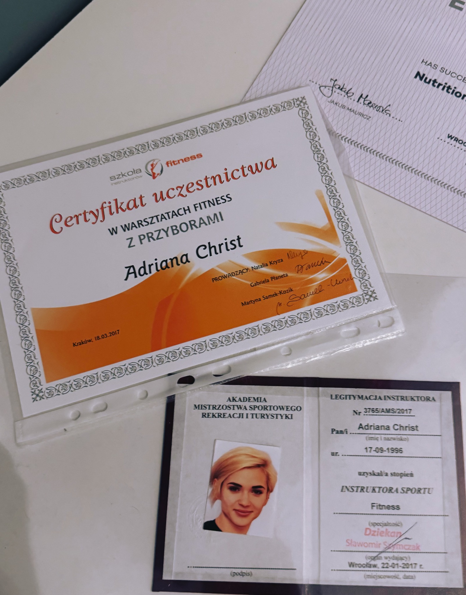 Certyfikat uczestnictwa w warsztatach fitness i legitymacja instruktora sportu Adriana Christ, leżące na jasnym tle, z widocznym podpisem i pieczęcią.