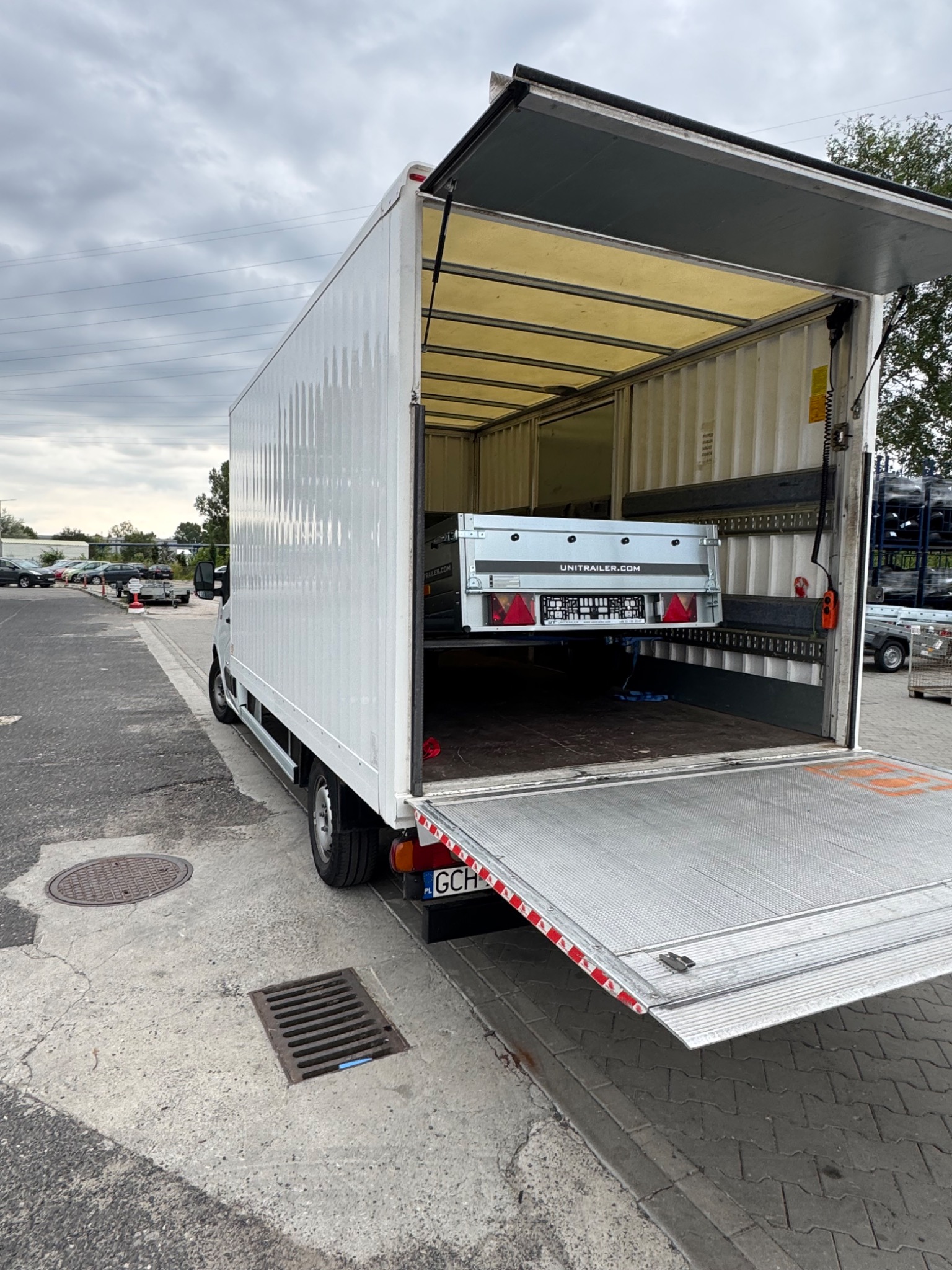 Biały samochód dostawczy z otwartą przestrzenią ładunkową, wewnątrz przyczepka. Widok z tyłu pojazdu na szarym tle parkingu.