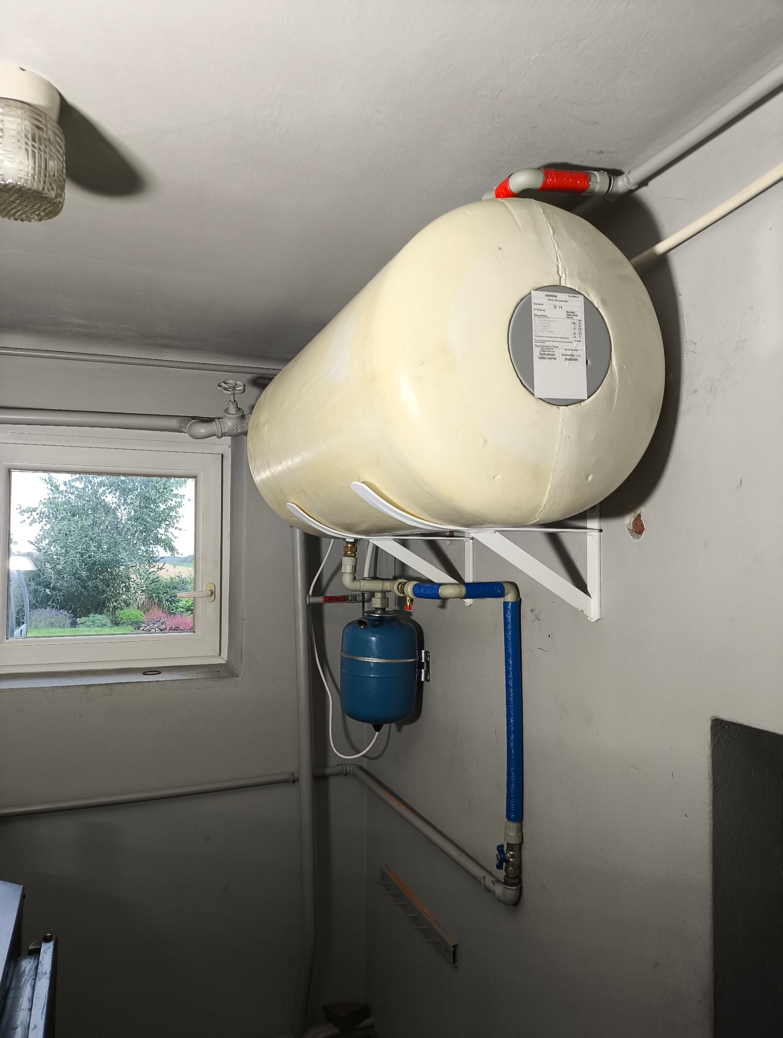 Bojler wiszący na ścianie z niebieskim zbiornikiem wyrównawczym i widocznymi rurami instalacji w szarym pomieszczeniu z oknem.