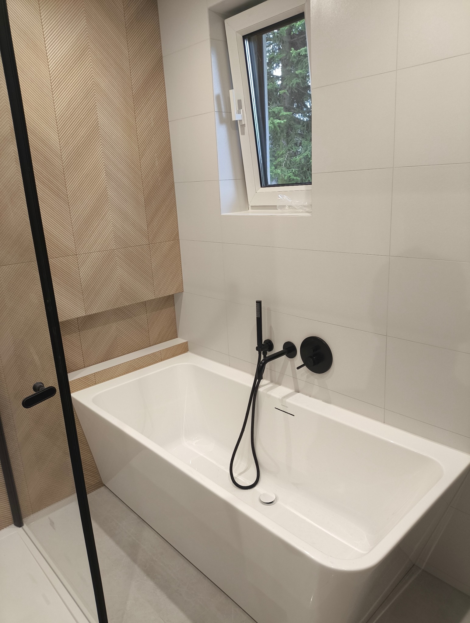Nowoczesna łazienka z białą wanną, czarną armaturą i dekoracyjną ścianą z drewnopodobnych płytek ułożonych w jodełkę. Okno z widokiem na zieleń.