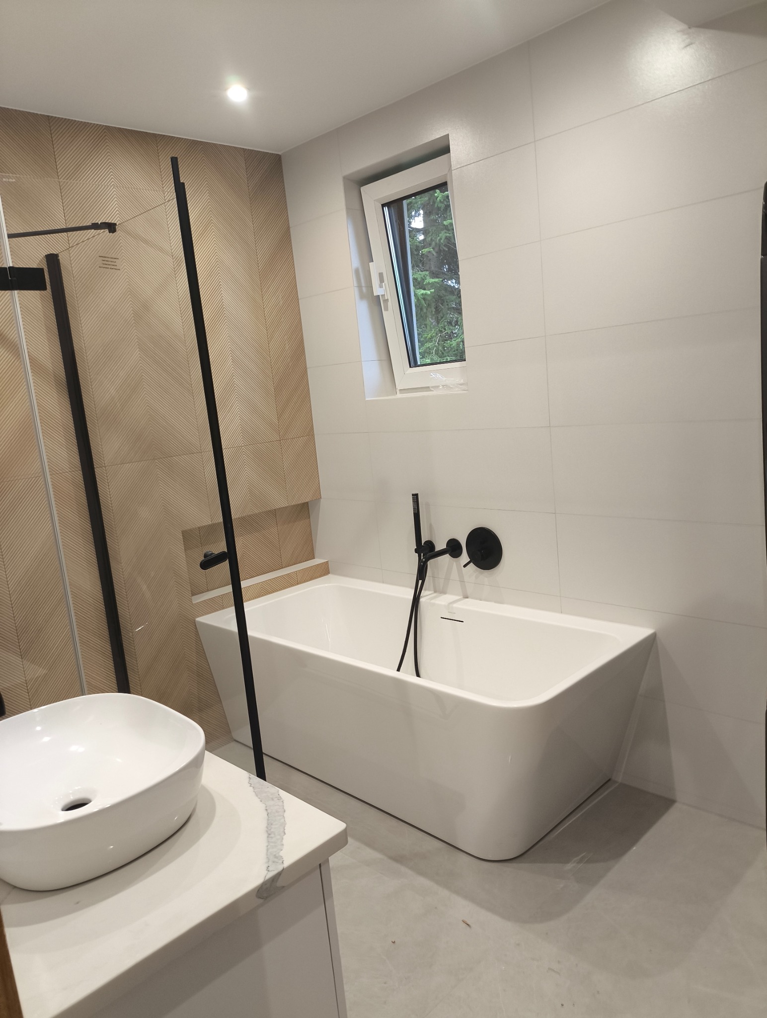 Nowoczesna łazienka z białą wanną wolnostojącą, czarną armaturą i umywalką nablatową na białym blacie. Ściana wyłożona płytkami imitującymi drewno.