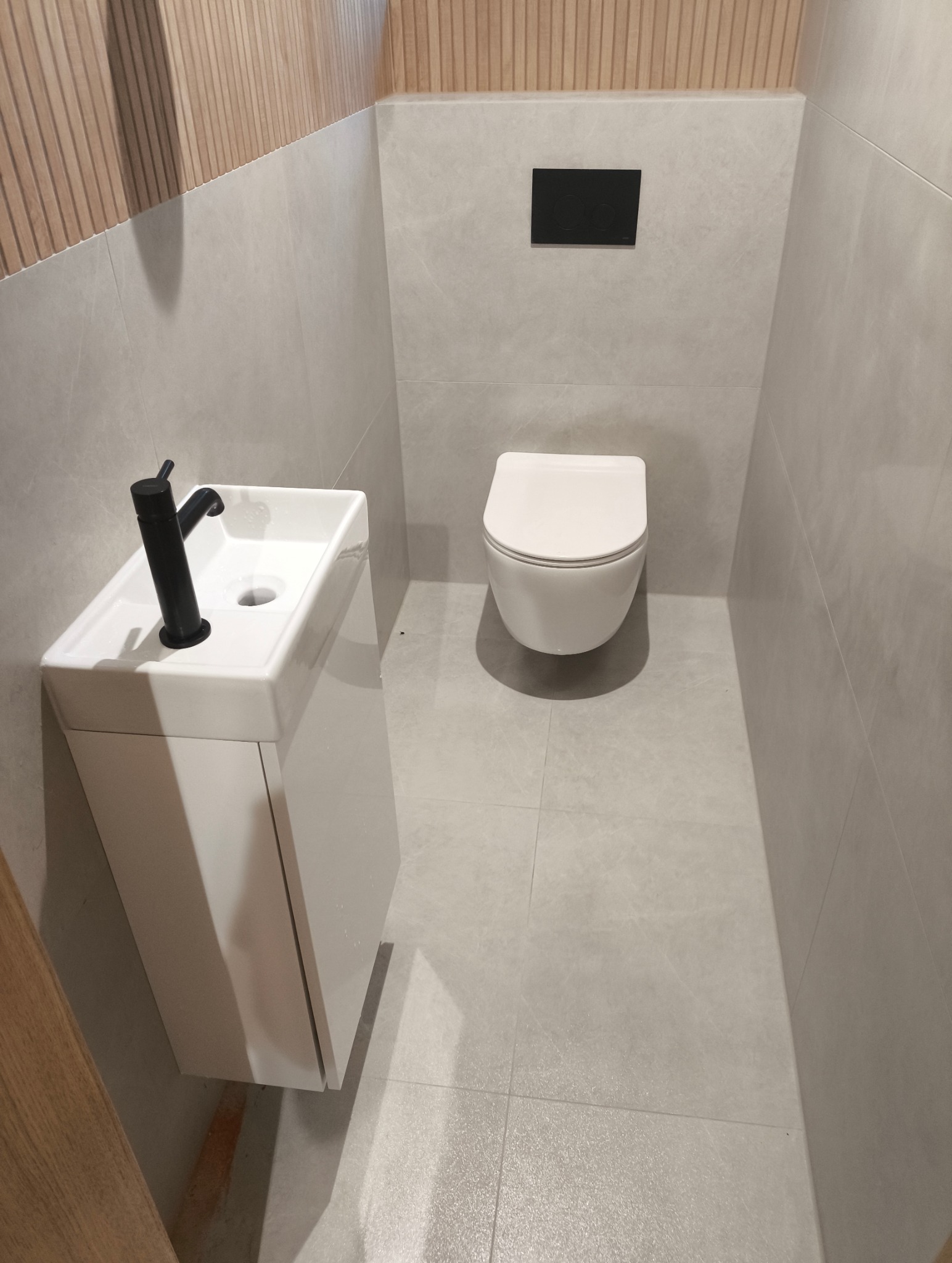 Nowoczesna łazienka z podwieszaną toaletą i umywalką z czarną armaturą na tle szarych płytek i drewnianej boazerii.