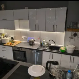 Nowoczesna kuchnia z białymi szafkami, czarnym blatem i podświetleniem LED. Na blacie stoją naczynia, czajnik i produkty spożywcze. Widoczne sprzęty AGD.