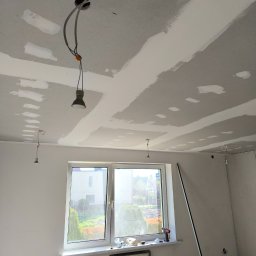DEBOR PROJECT SPÓŁKA Z OGRANICZONĄ ODPOWIEDZIALNOŚCIĄ - Pokój w trakcie remontu: sufit z płyt karton-gips, widoczne łączenia szpachlowane, wiszące żarówki, okno z widokiem na zieleń, narzędzia na parapecie.