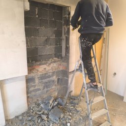 DEBOR PROJECT SPÓŁKA Z OGRANICZONĄ ODPOWIEDZIALNOŚCIĄ - Pracownik na drabinie w Goczałkowicach-Zdroju, wykuwa otwór w ścianie z szarej cegły. Gruz na podłodze. Widoczne fragmenty instalacji i płyty karton-gips.