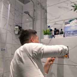 DEBOR PROJECT SPÓŁKA Z OGRANICZONĄ ODPOWIEDZIALNOŚCIĄ - Instalacja rewizji w łazience z marmurowymi płytkami. Mężczyzna w białej koszulce używa pomarańczowego śrubokręta. Widoczna kabina prysznicowa i półka z kosmetykami.