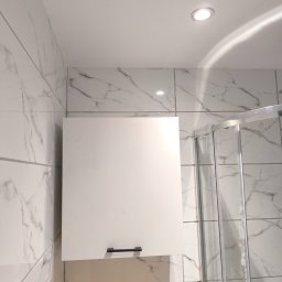 DEBOR PROJECT SPÓŁKA Z OGRANICZONĄ ODPOWIEDZIALNOŚCIĄ - Biała szafka łazienkowa z czarnym uchwytem, marmurowe płytki na ścianach i fragment kabiny prysznicowej. Nowoczesne wykończenie wnętrza łazienki.