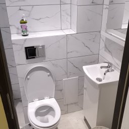 DEBOR PROJECT SPÓŁKA Z OGRANICZONĄ ODPOWIEDZIALNOŚCIĄ - Biała łazienka po remoncie: marmurowe płytki, podwieszana toaleta z chromowanym przyciskiem, umywalka z szafką i lustro. Na półce butelka płynu do czyszczenia.