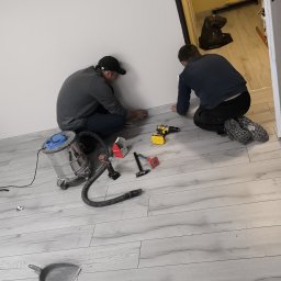 DEBOR PROJECT SPÓŁKA Z OGRANICZONĄ ODPOWIEDZIALNOŚCIĄ - Montaż paneli podłogowych: dwóch monterów przy pracy, narzędzia (wiertarka, młotek, odkurzacz) rozłożone na jasnej podłodze imitującej drewno, białe ściany.