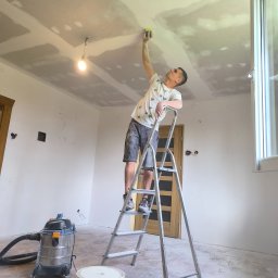 DEBOR PROJECT SPÓŁKA Z OGRANICZONĄ ODPOWIEDZIALNOŚCIĄ - Mężczyzna na drabinie szlifuje sufit z płyt gipsowo-kartonowych w jasnym wnętrzu. Widoczny odkurzacz przemysłowy i prowizoryczne oświetlenie.