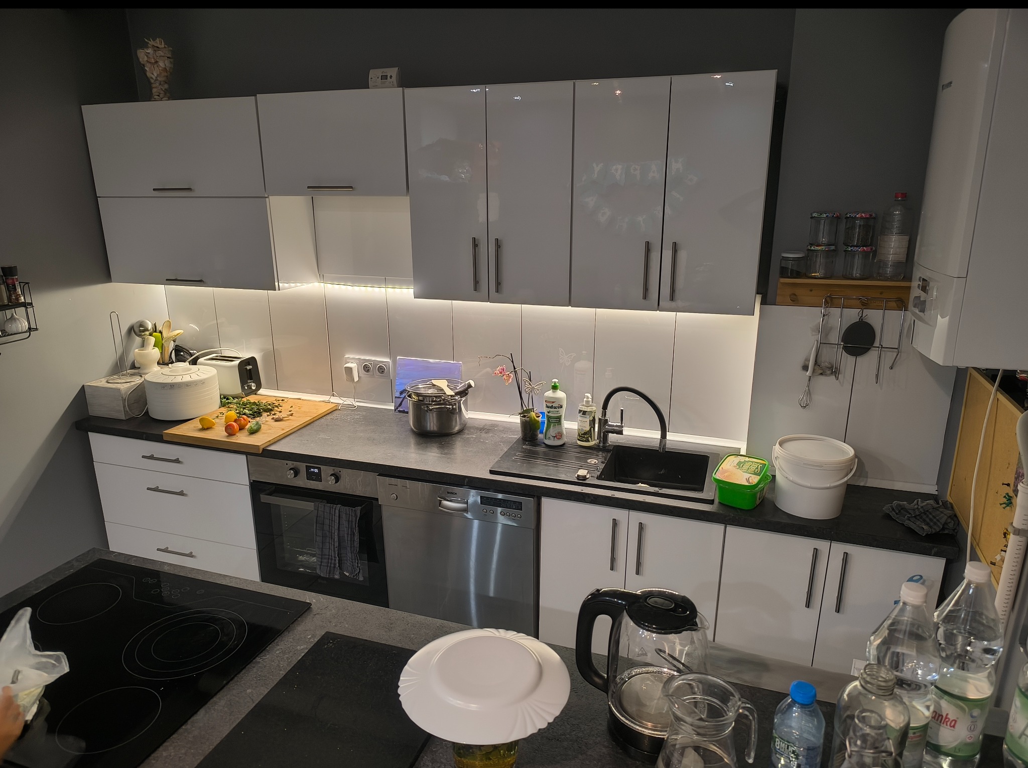 Nowoczesna kuchnia z białymi szafkami, czarnym blatem i podświetleniem LED. Na blacie stoją naczynia, czajnik i produkty spożywcze. Widoczne sprzęty AGD.