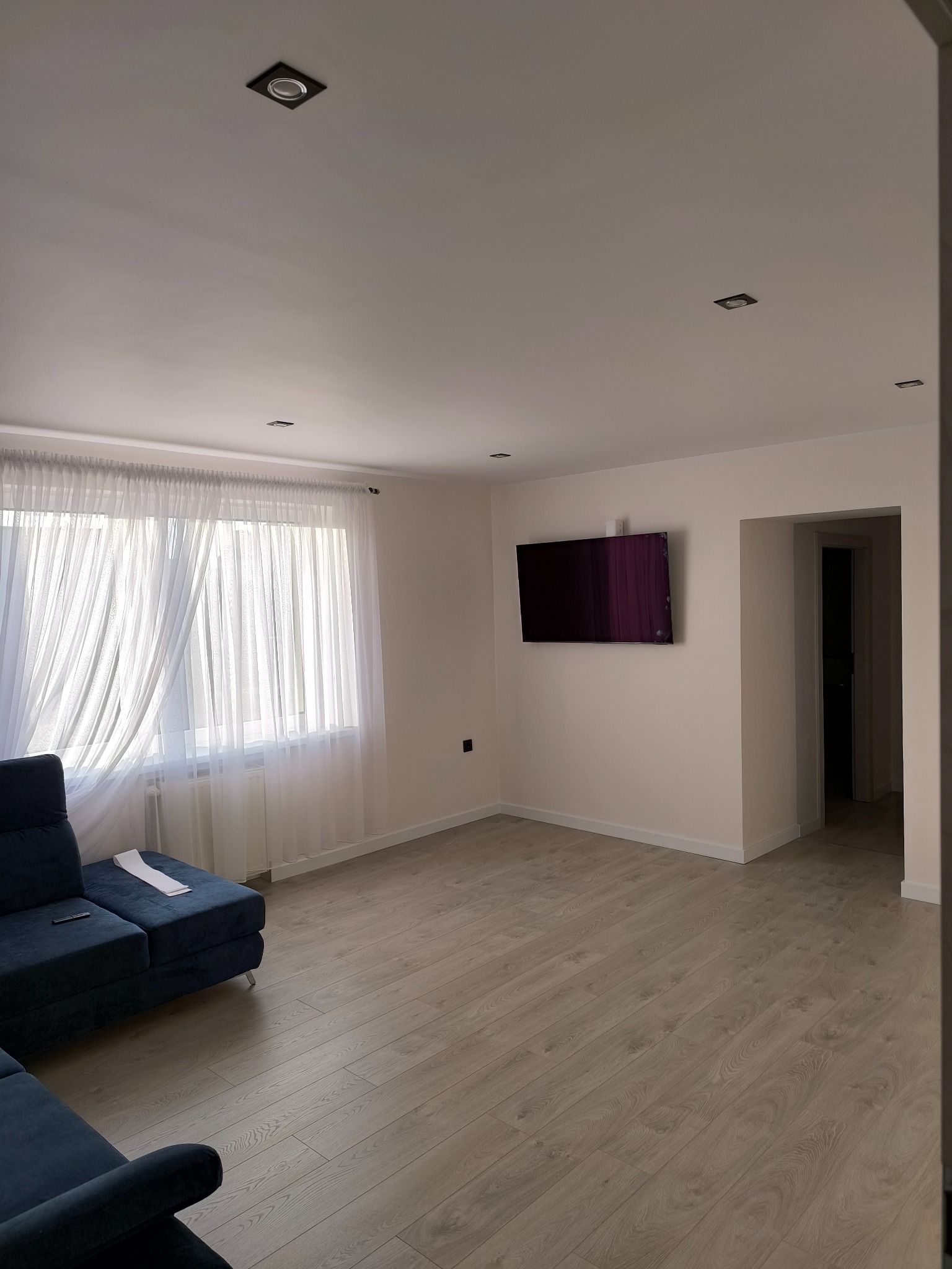 Jasne, przestronne wnętrze po remoncie: panele podłogowe, ściany w odcieniach bieli, okno z firanami i telewizor na ścianie. Minimalistyczny design z nowoczesnym oświetleniem.