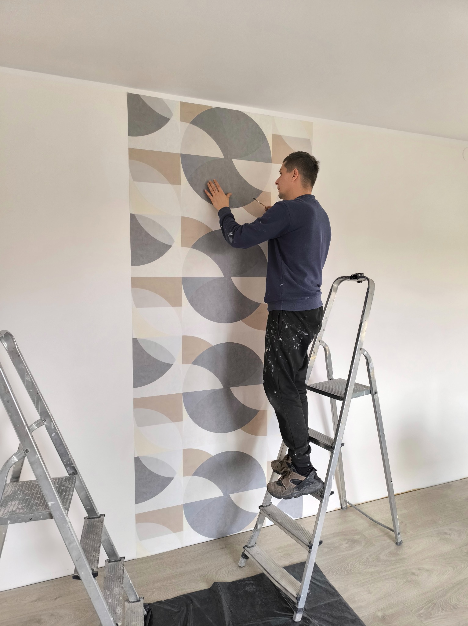 Mężczyzna na drabinie aplikuje tapetę w geometryczne wzory na ścianie. Widoczne narzędzia i zabezpieczenia podłogi. Jasne wnętrze w trakcie wykończenia.