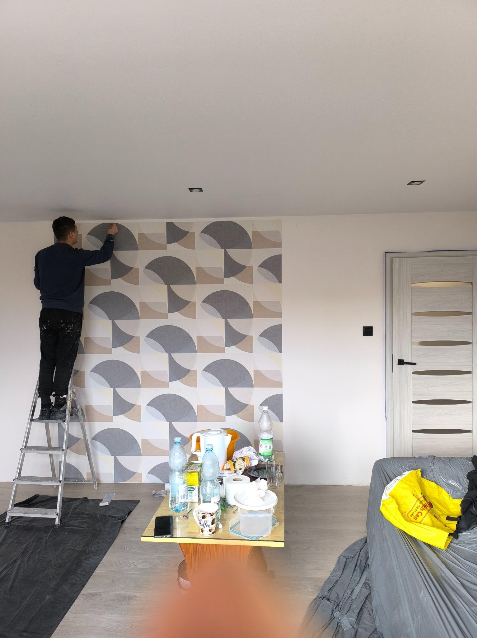 Mężczyzna na drabinie aplikuje tapetę w geometryczne wzory na ścianie. Widoczne narzędzia i zabezpieczone meble. Częściowo wykończone pomieszczenie.