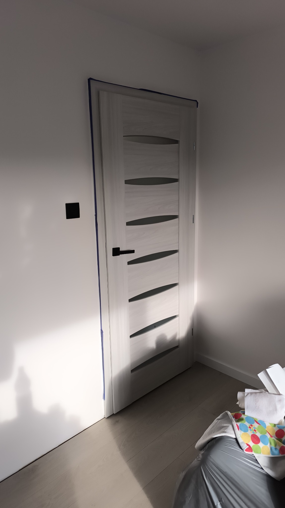 Białe drzwi wewnętrzne z poziomymi przeszkleniami, zabezpieczone taśmą malarską przy ościeżnicy, po malowaniu ściany na biało. Widoczny fragment podłogi i ściany.