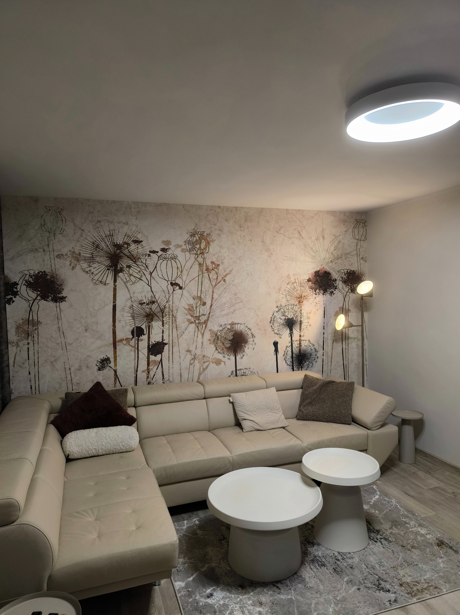 Salon z narożnikiem w odcieniach beżu, stolikami kawowymi i tapetą w motywy roślinne. Nowoczesne oświetlenie sufitowe i podłogowa lampa dopełniają aranżację.