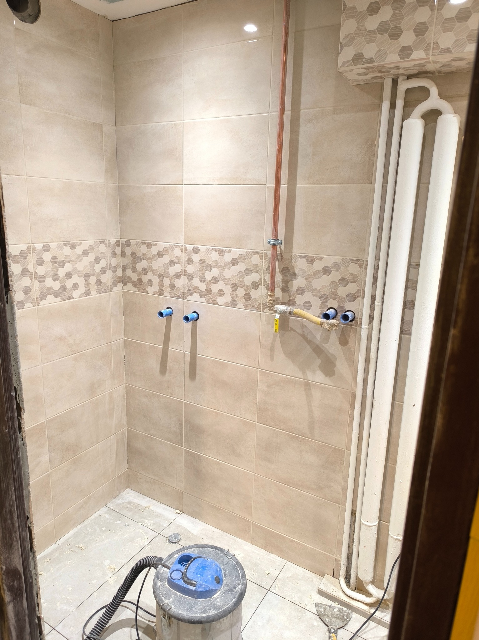 Wnętrze łazienki w trakcie remontu: beżowe płytki z dekoracyjnym pasem, widoczne rury instalacji wodnej i grzejnik, odkurzacz przemysłowy na podłodze.