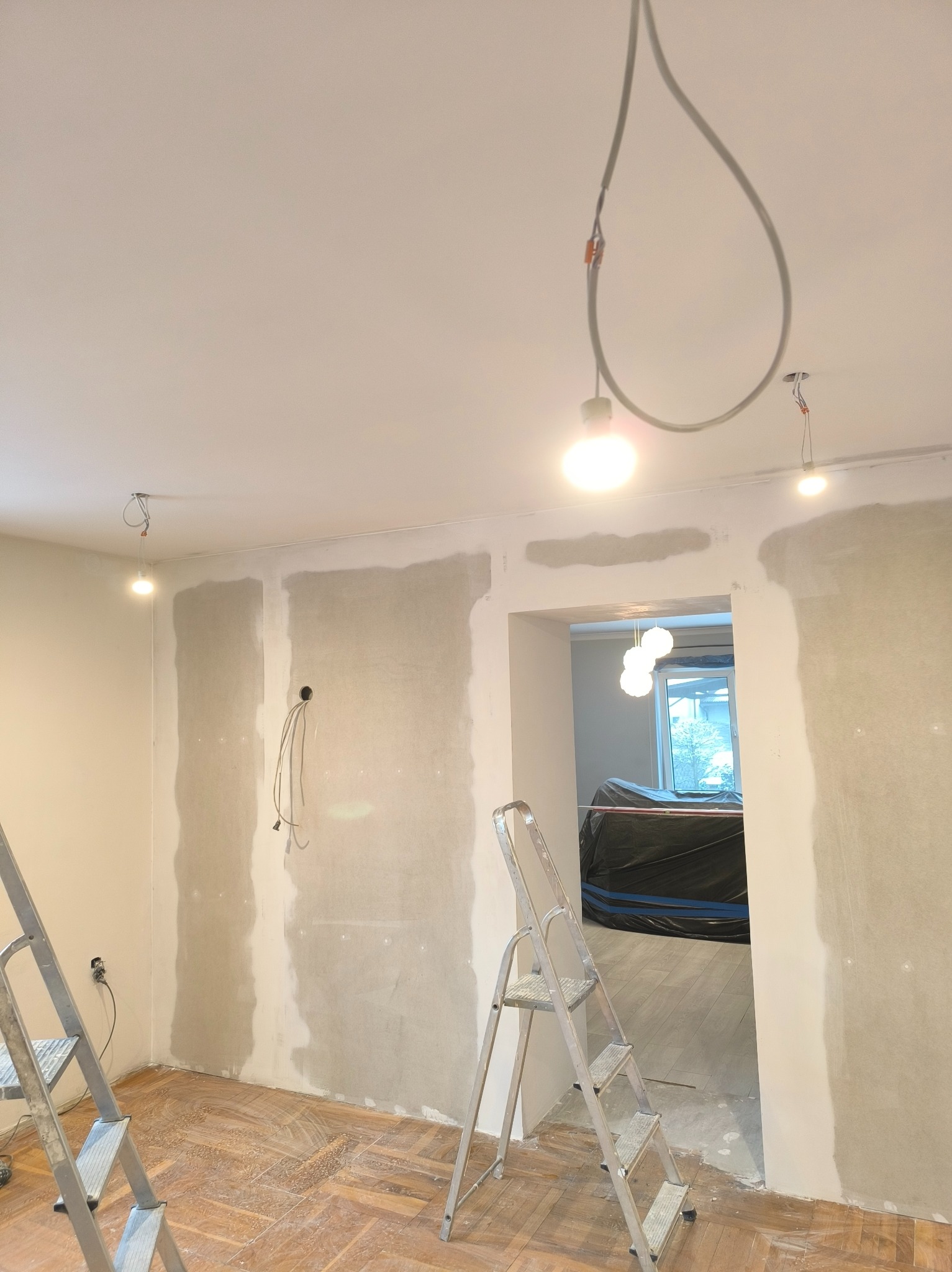 Pokój w trakcie szpachlowania ścian, widoczne drabiny, wiszące kable oświetleniowe i fragment parkietu. Przez otwór widać zabezpieczone folią meble.