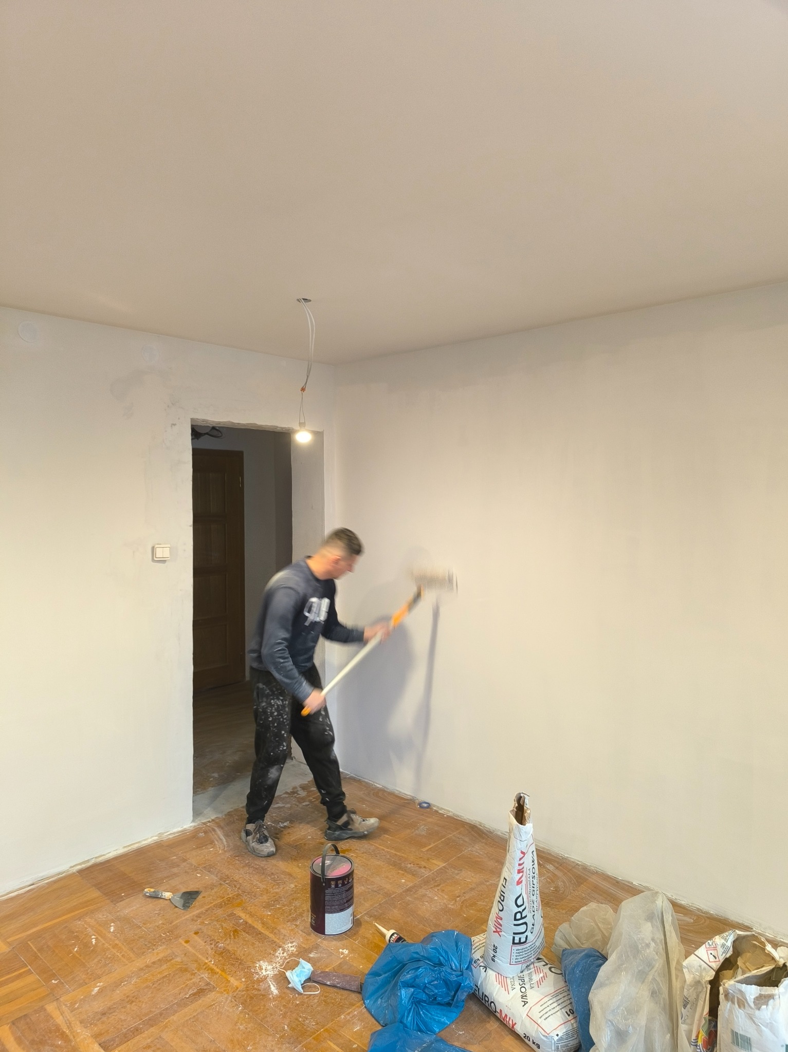 Mężczyzna maluje ścianę wałkiem w pokoju z parkietem. Widoczne narzędzia malarskie, puszka farby i worki z gipsem. Ściana świeżo pomalowana na jasny kolor.