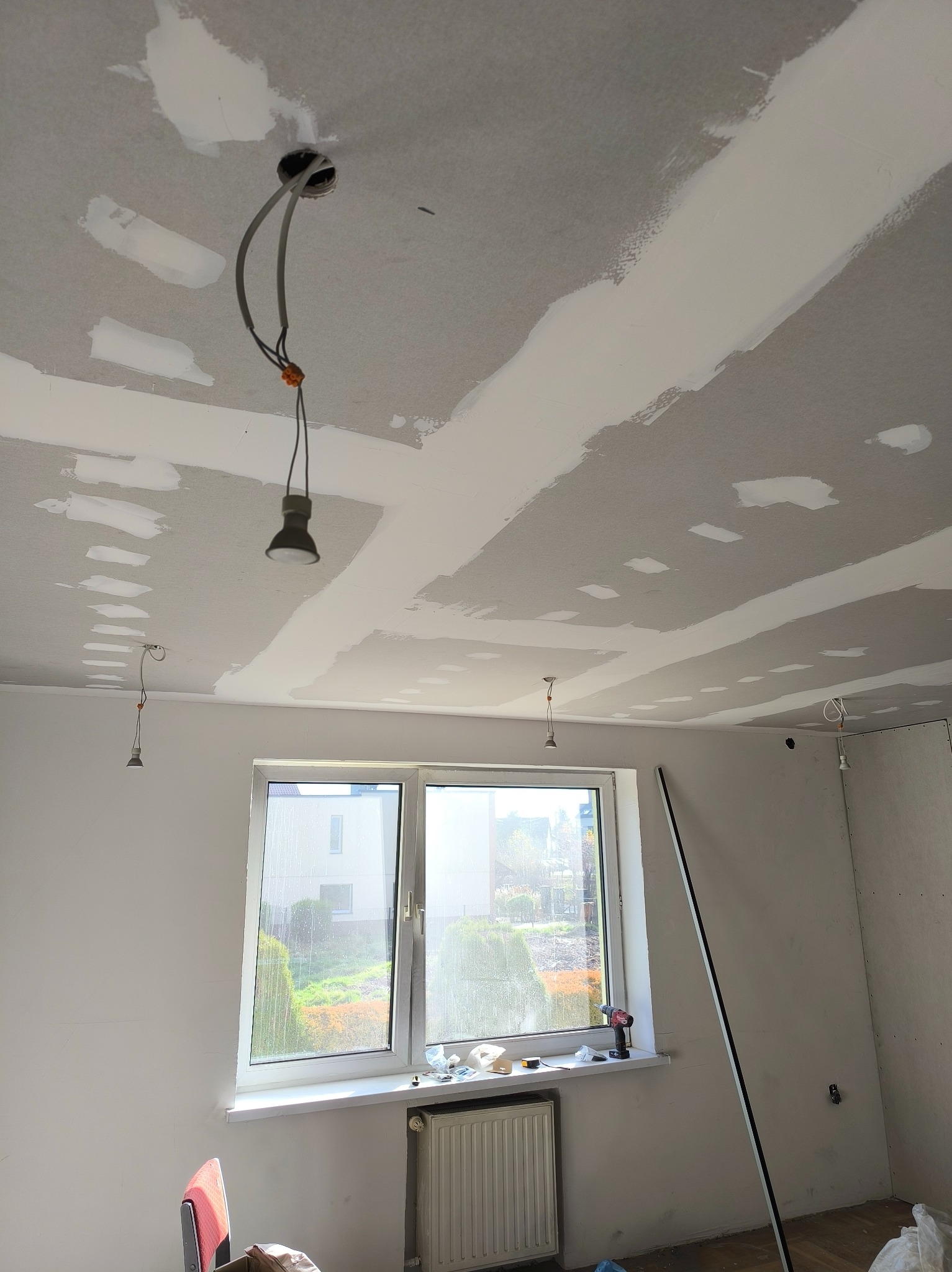 Pokój w trakcie remontu: sufit z płyt karton-gips, widoczne łączenia szpachlowane, wiszące żarówki, okno z widokiem na zieleń, narzędzia na parapecie.