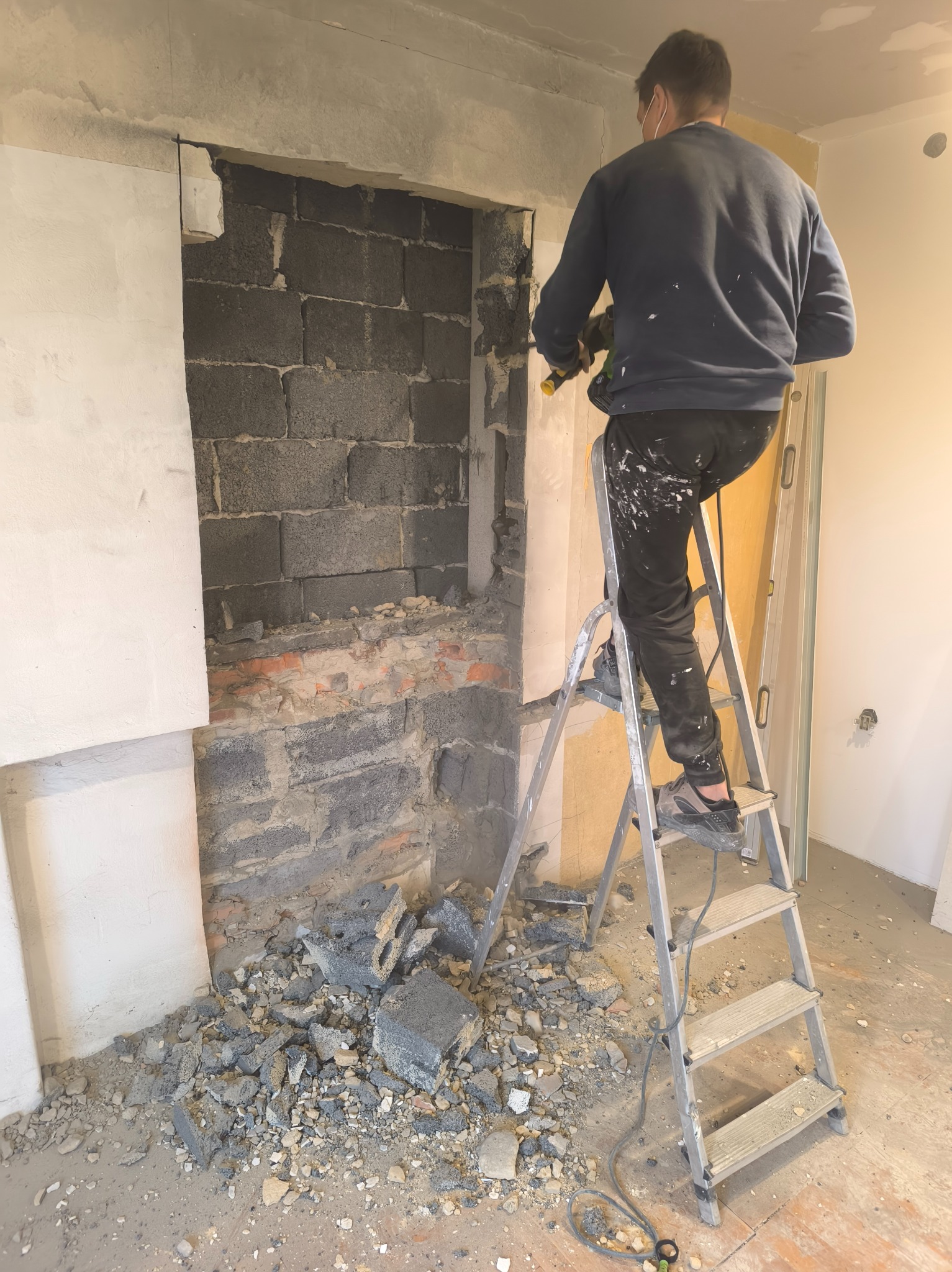 Pracownik na drabinie w Goczałkowicach-Zdroju, wykuwa otwór w ścianie z szarej cegły. Gruz na podłodze. Widoczne fragmenty instalacji i płyty karton-gips.