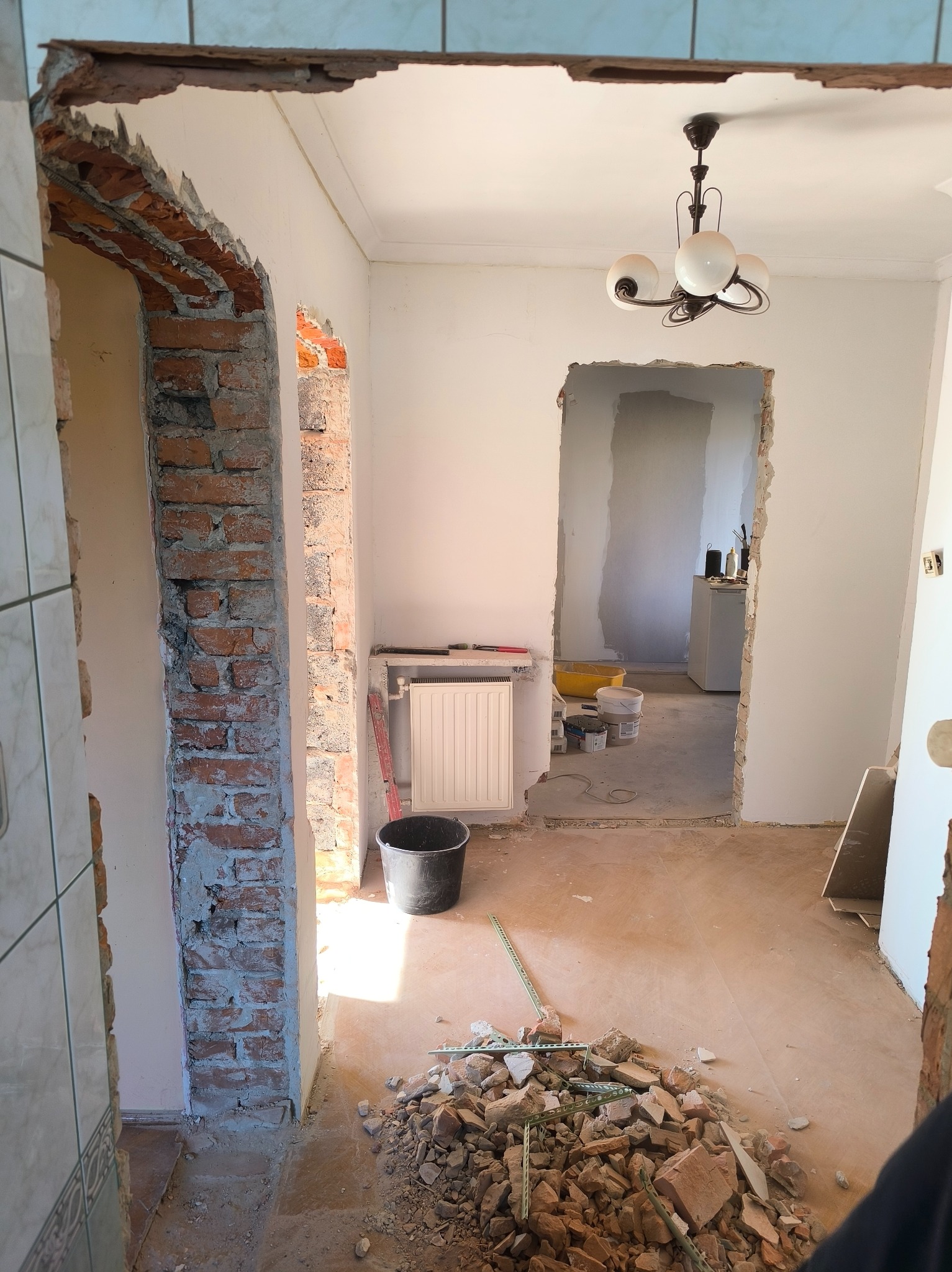 Demontaż ścian działowych w mieszkaniu: widoczne cegły, gruz, przygotowanie do remontu. W tle grzejnik i lampa sufitowa. Ujęcie z perspektywy korytarza.