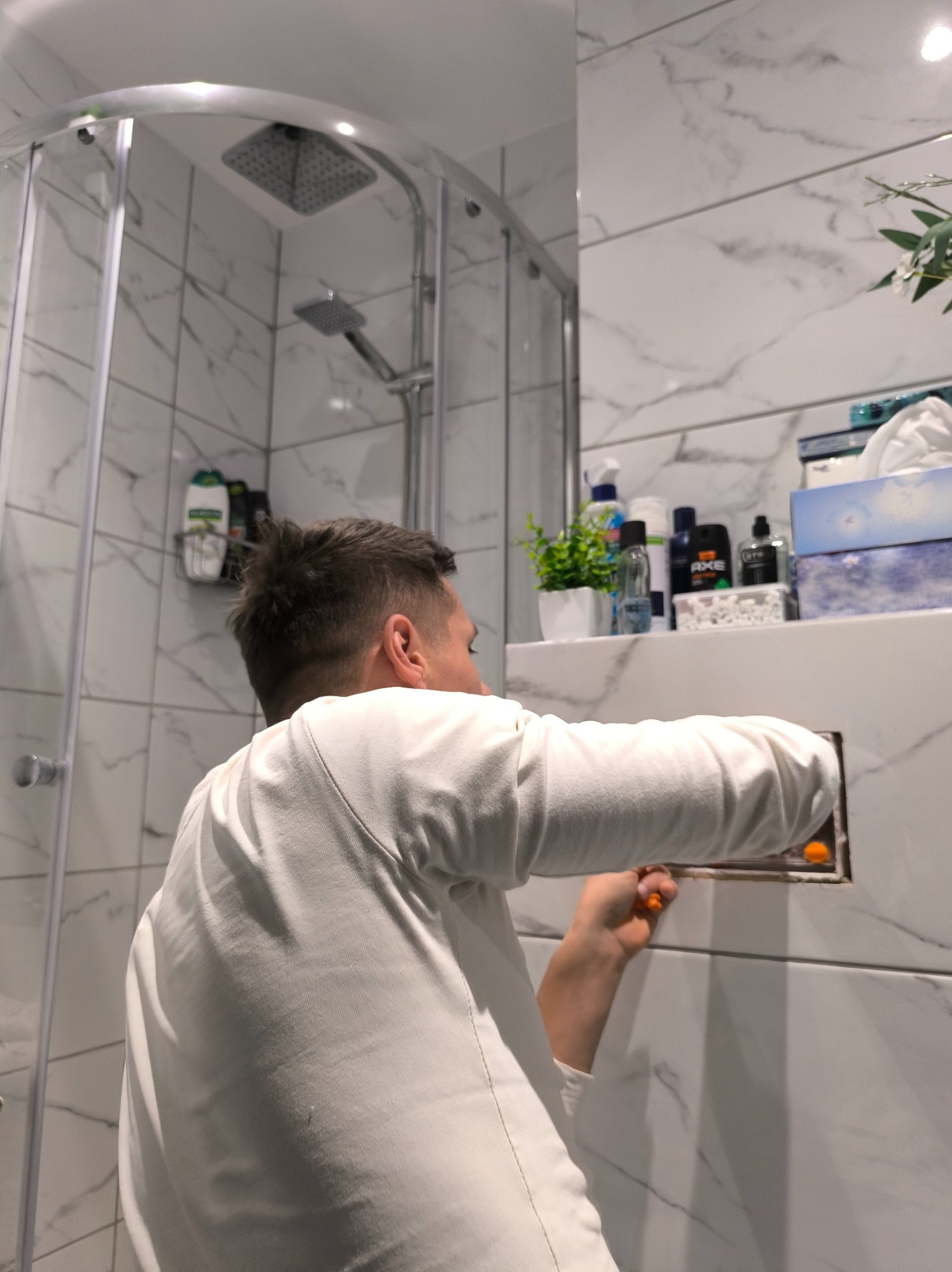 Instalacja rewizji w łazience z marmurowymi płytkami. Mężczyzna w białej koszulce używa pomarańczowego śrubokręta. Widoczna kabina prysznicowa i półka z kosmetykami.