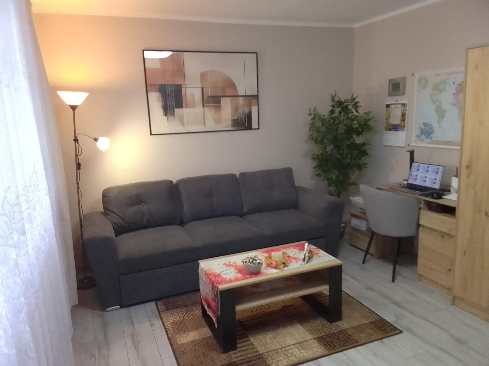 Szary narożnik w salonie z obrazem i lampą. Na stoliku dekoracje. Widok na biurko z laptopem i mapą świata. Jasne ściany i podłoga.