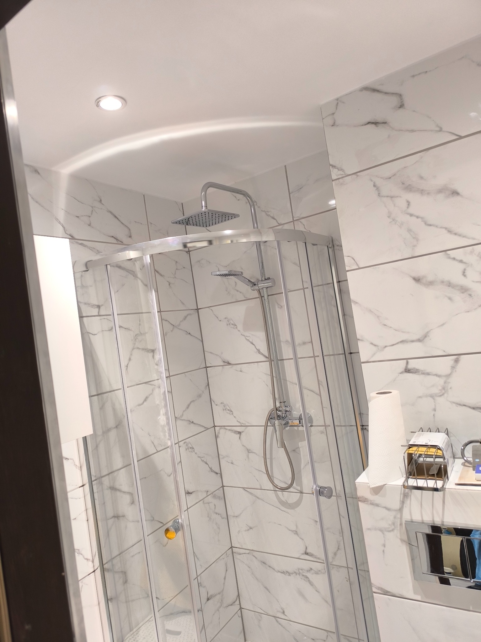 Nowoczesna kabina prysznicowa z deszczownicą i marmurowymi płytkami. Chromowane wykończenia, minimalistyczny design. Widok na detale armatury i czystą, jasną przestrzeń.