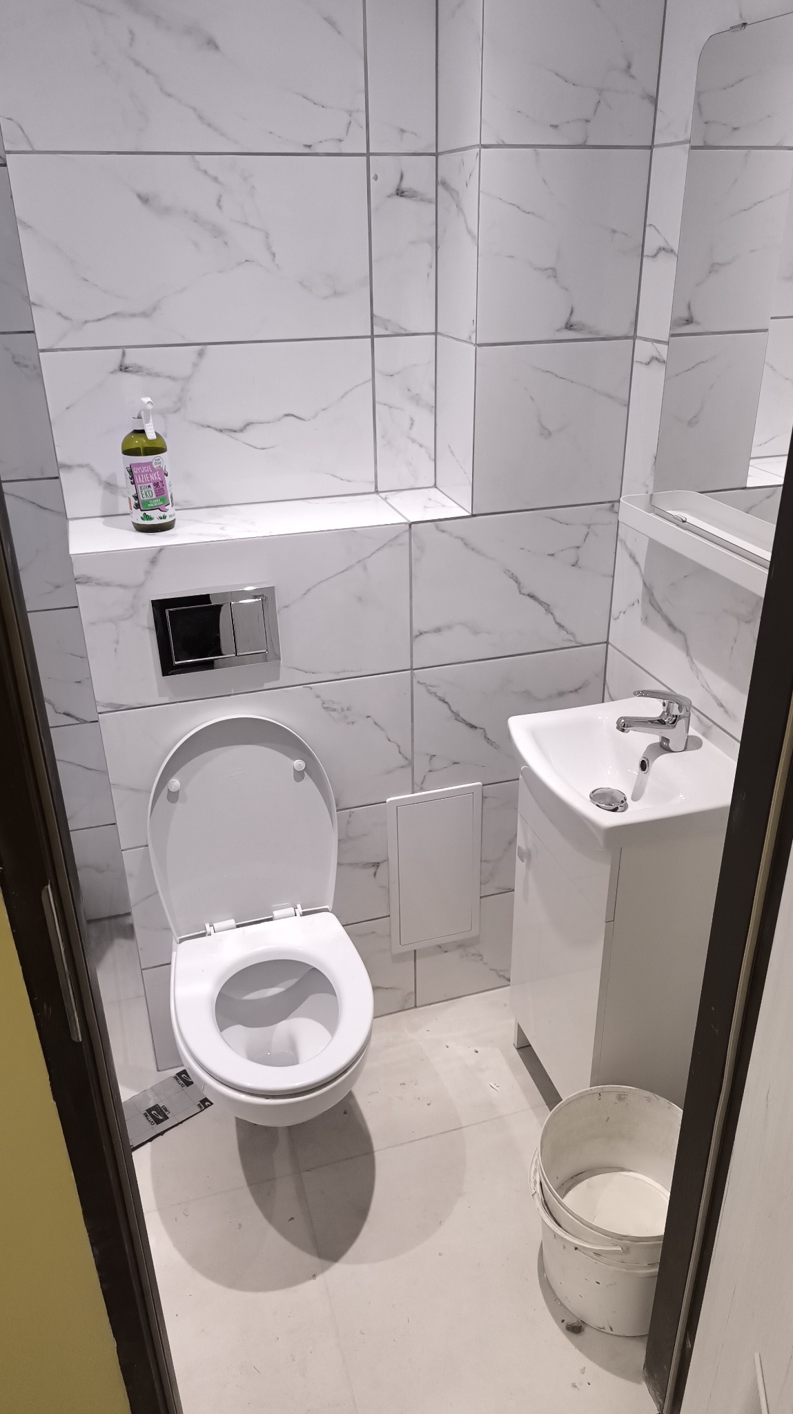 Biała łazienka po remoncie: marmurowe płytki, podwieszana toaleta z chromowanym przyciskiem, umywalka z szafką i lustro. Na półce butelka płynu do czyszczenia.
