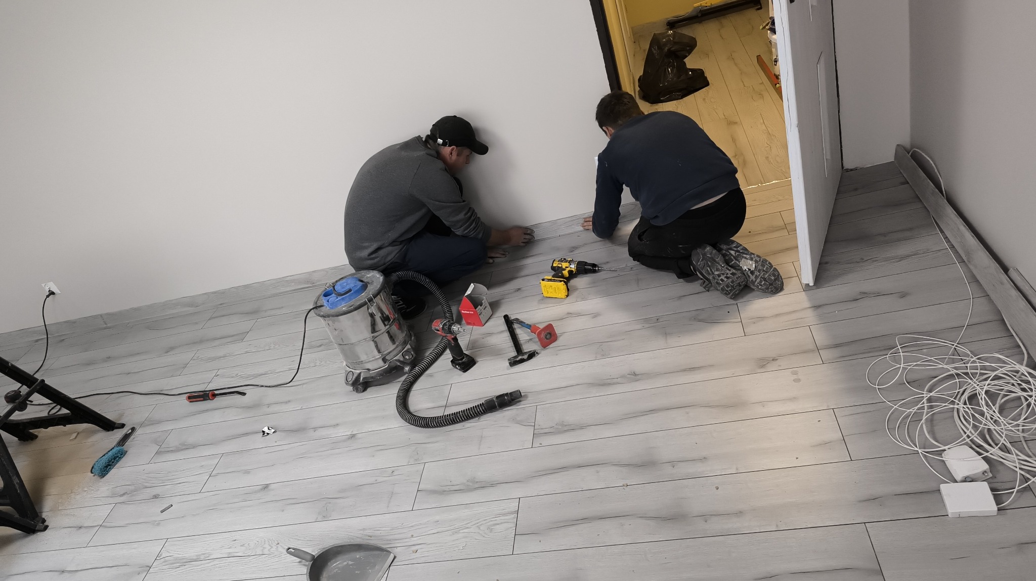 Montaż paneli podłogowych: dwóch monterów przy pracy, narzędzia (wiertarka, młotek, odkurzacz) rozłożone na jasnej podłodze imitującej drewno, białe ściany.