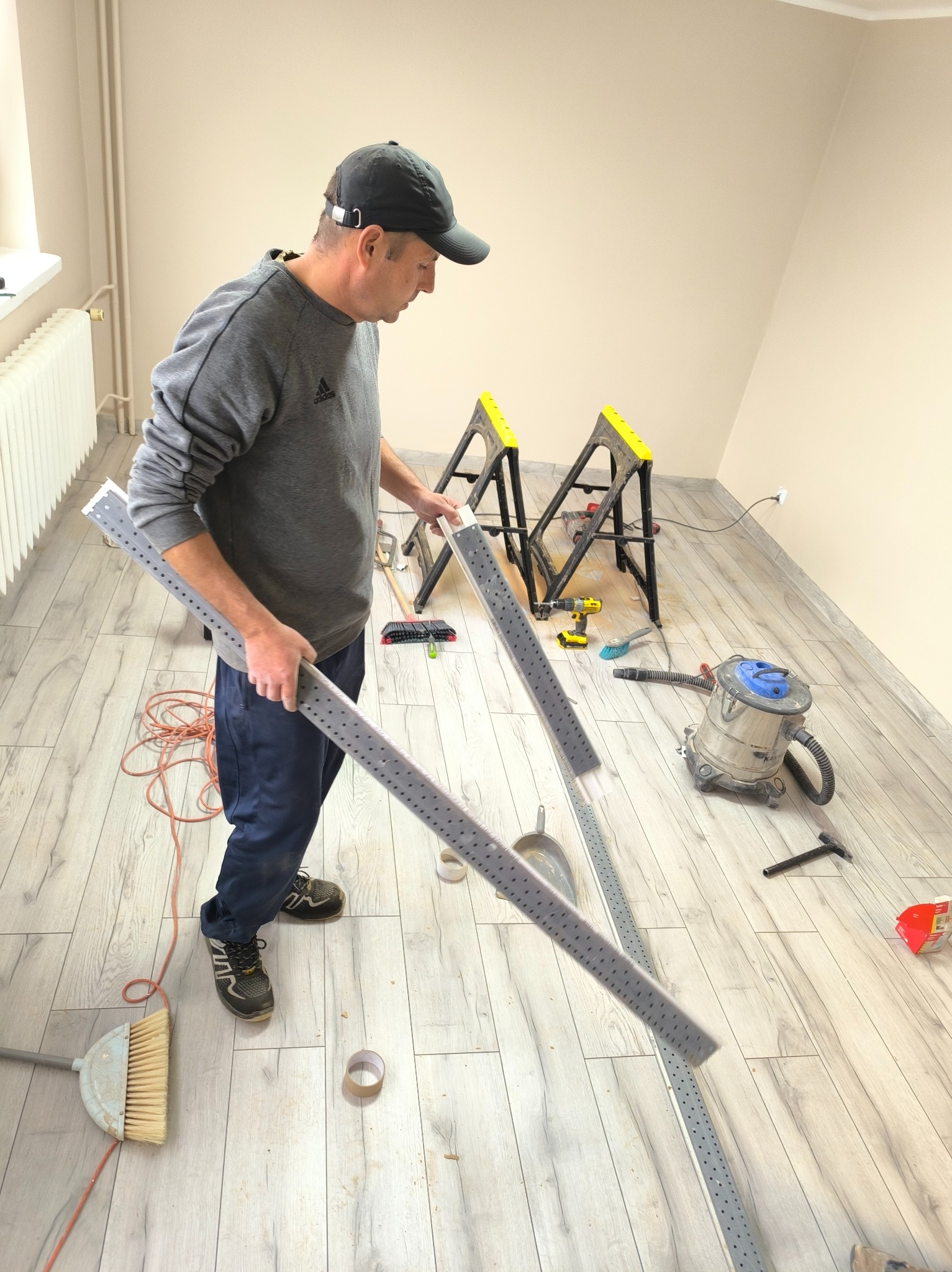 Instalator trzyma aluminiowe profile z otworami na tle podłogi z paneli i narzędzi, przygotowując się do montażu. Widoczne kozły, odkurzacz i szczotka.