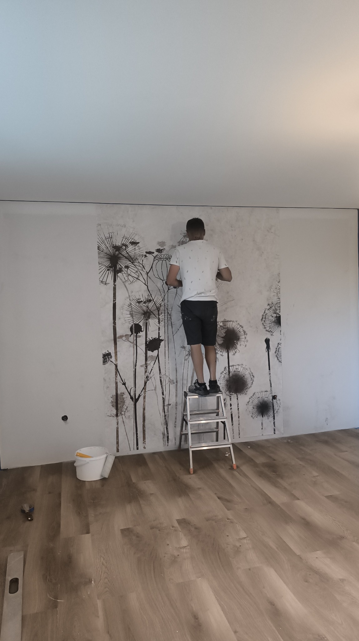 Mężczyzna na drabinie tapetuje ścianę w pokoju. Widoczny wzór dmuchawców na tapecie, wiadro z klejem i narzędzia na podłodze. Częściowo wykończona ściana.