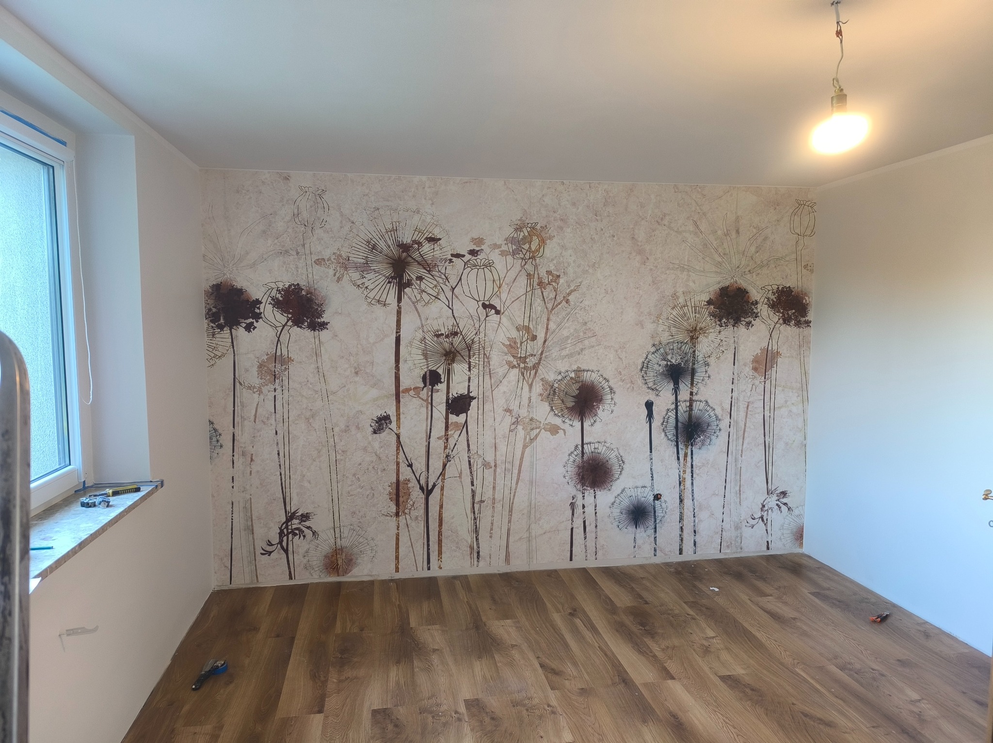 Pokój po tapetowaniu z motywem dmuchawców na ścianie, drewniana podłoga, okno z lewej strony. Widoczne narzędzia na parapecie i podłodze.