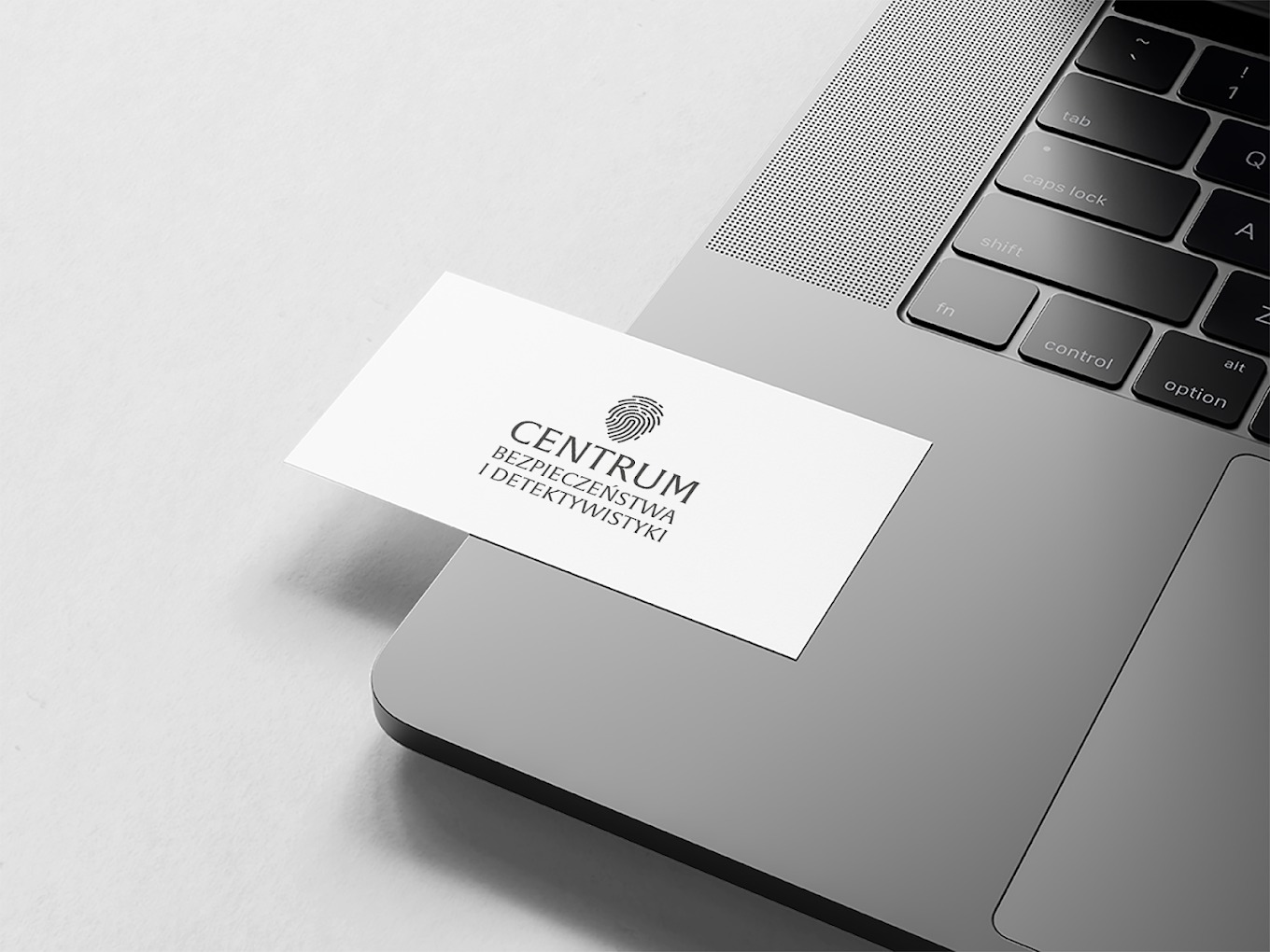 Wizytówka 'Centrum Bezpieczeństwa i Detektywistyki' leży na szarym laptopie, ukazując logo z odciskiem palca i czcionkę bezszeryfową. Minimalistyczny design.